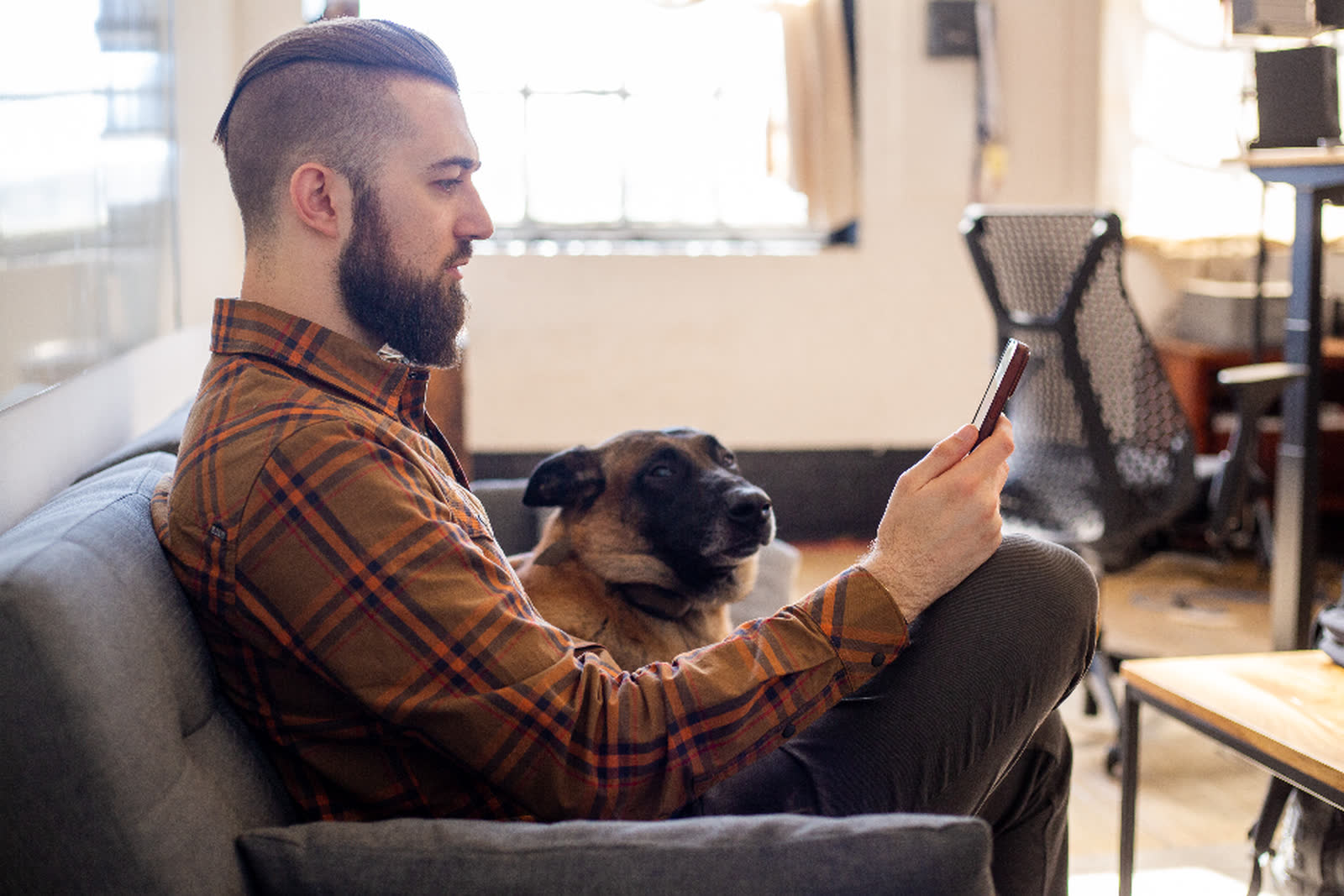 A man sitting on a sofa scrolling on his phone, with his dog beside him