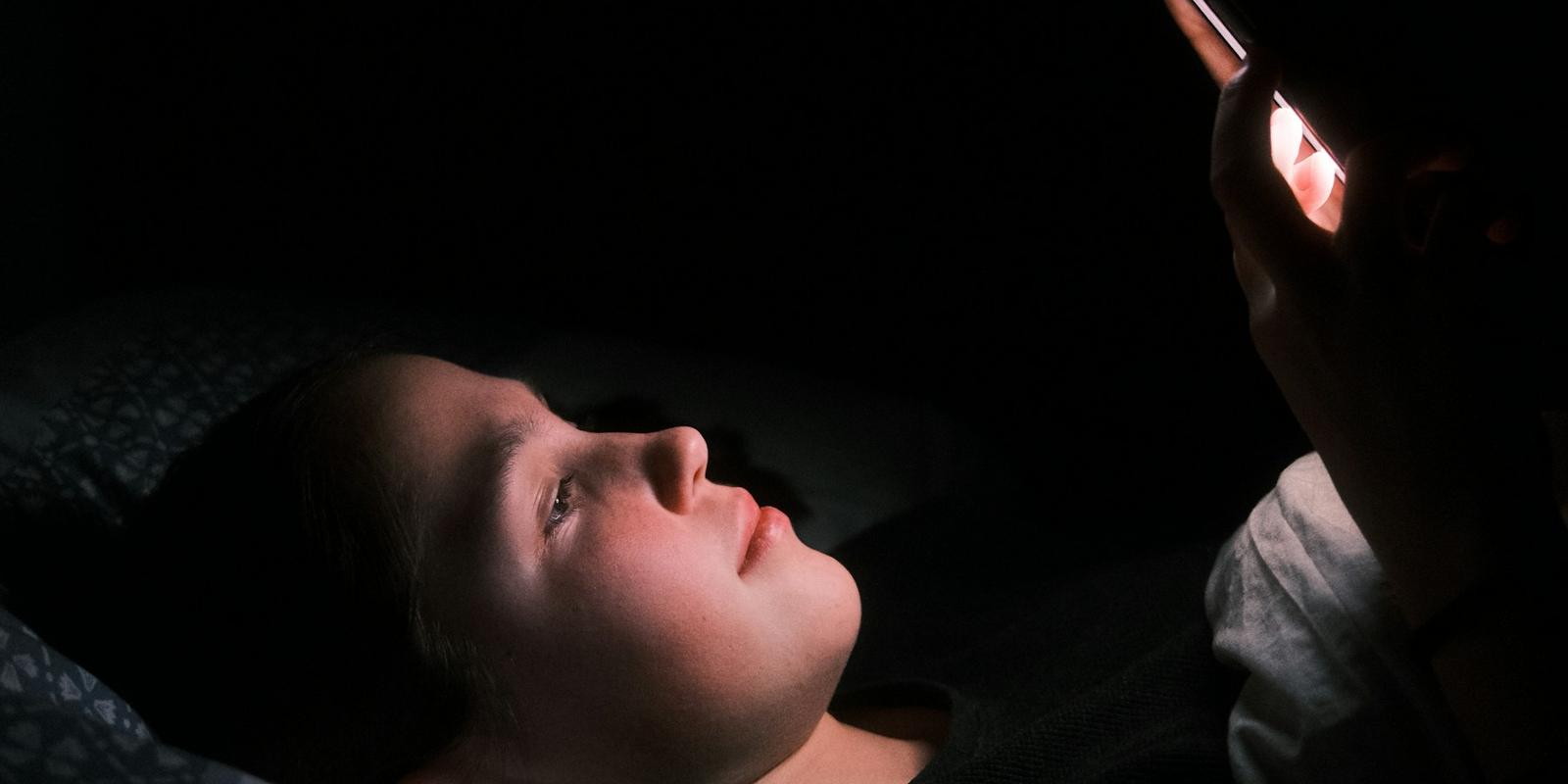 A person lying in a dark bedroom, face lit by a phone screen